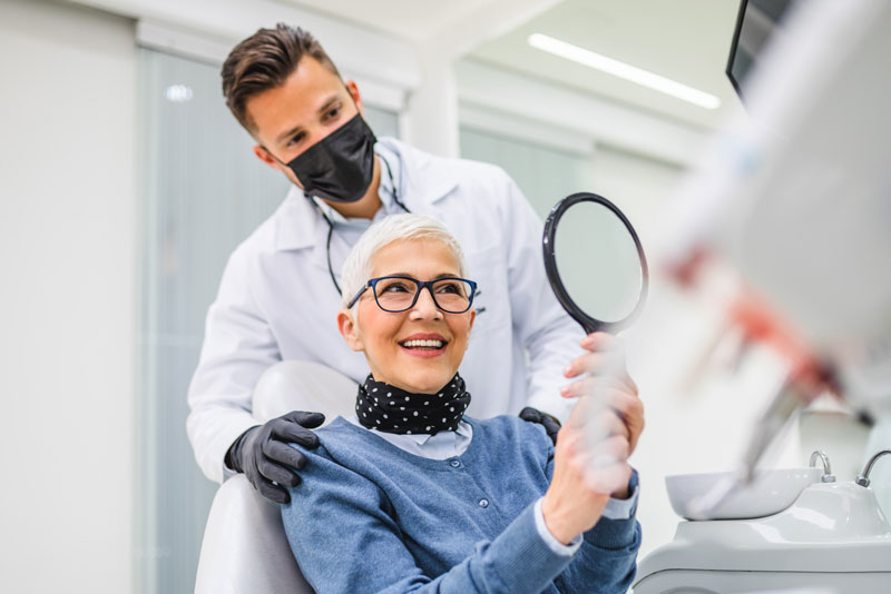 a picture of a dental patient and her doctor smiling at the patient through a hand mirror because she is now treated with CeraRoot dental implants