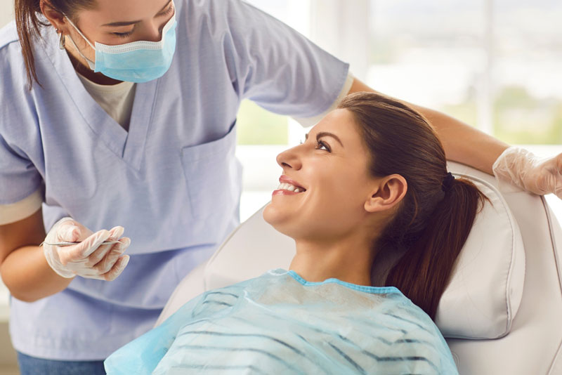 A dentist wearing a mask and gloves talking to a smiling female patient in a dental chair during an appointment.