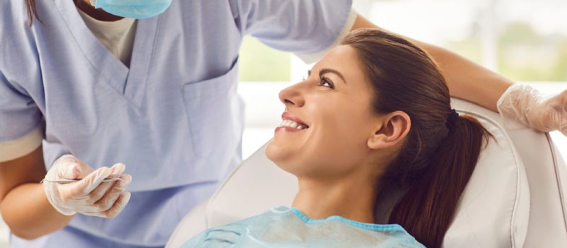 A dentist wearing a mask and gloves talking to a smiling female patient in a dental chair during an appointment.