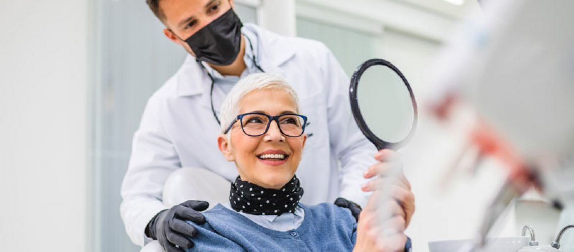 a picture of a dental patient and her doctor smiling at the patient through a hand mirror because she is now treated with CeraRoot dental implants